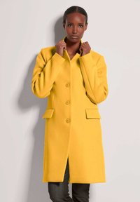 Geel wollen mengseljas met een geknipt kraag, vier knopen en twee voorzakken. De textuur is glad, met een getailleerde pasvorm.