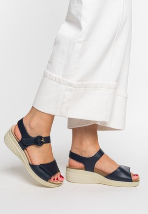 Navy leather wedge sandals with a rounded open toe, ankle strap, and circular buckle detail. Light-colored platform sole.