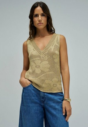 Mujer con cabello largo y oscuro, vestida con un top sin mangas beige de punto con patrones florales y jeans azules holgados, de pie con una mano en el bolsillo.