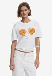 Hvid T-shirt med kort ærme, der har orange skjellgrafik og tekst. Bæres med højtkalibrerede hvide bukser, der har en glat tekstur og en afslappet pasform.