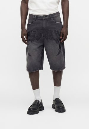 Mann trägt dunkelgraue knielange Jeansshorts mit schwarzem grafischem Design, weiße Socken, schwarze Loafers und ein weißes T-Shirt, stehend vor weißem Hintergrund.