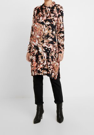 Femme portant une tunique boutonnée à manches longues, longueur genou, avec un motif abstrait noir, marron et beige, un pantalon noir et des bottes noires à talons.