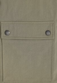 Khakifarbenes Material mit glatter Textur, das über eine Fronttasche mit Klappe verfügt, die mit zwei silbernen Druckknöpfen gesichert ist. Minimalistisches Design mit unauffälligen Nähten.