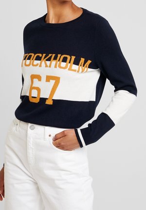 Person iført en marineblå, hvid og guldfarvet sweater med teksten "STOCKHOLM 67", kombineret med højtaljede hvide bukser, hånd i lommen.
