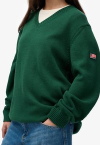 Groene v-halstrui met ribgebreide manchetten en zoom, gemaakt van een textuur gebreid materiaal. Voorzien van een klein label met "Superdry" en een vlagontwerp.