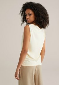 Light yellow sleeveless top with a slight drape, paired with beige, textured wide-leg trousers. Smooth fabric with minimal detailing.