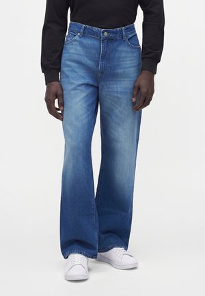 Mann mit blauen Jeans mit weitem Bein, schwarzem Langarmshirt und weißen Sneakers steht vor einem einfarbigen Hintergrund.
