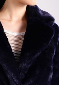 Abrigo de pelo sintético en navy con una textura suave y aterciopelada, que presenta un cuello con notch y se muestra sobre una blusa de malla blanca con escote redondo.