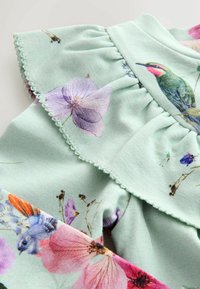 Mintgrüner Stoff mit einem floralen Muster, das rosa und lila Blumen sowie einen Kolibri zeigt, abgeschlossen mit einem Rüschensaum und gewelltem Besatz.