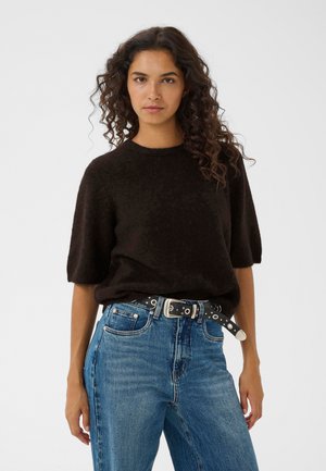 Ung kvinde med krøllet hår iført en sort kortærmet sweater, blå jeans og et sort bælte med sølvfarvede øjer og spænde.