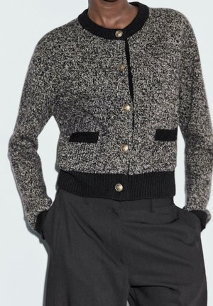 Personne portant un cardigan tricoté noir et blanc avec des boutons dorés et un pantalon noir, mains dans les poches, torse visible sur un fond uni.