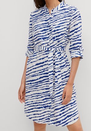 Mujer con un vestido camisero blanco hasta la rodilla con rayas horizontales azules que parecen pinceladas y un cinturón de tela a juego atado a la cintura.