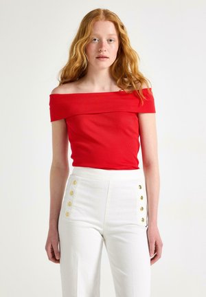 Donna con lunghi capelli rossi che indossa un top rosso scollato e pantaloni bianchi a vita alta con bottoni dorati, in piedi davanti a uno sfondo bianco.