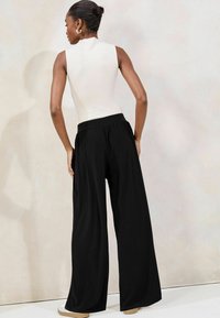 Witte mouwloze top in combinatie met zwarte wijde broek. De broek heeft een elastische tailleband en een gladde, vloeiende stof, afgemaakt met lichte schoenen.