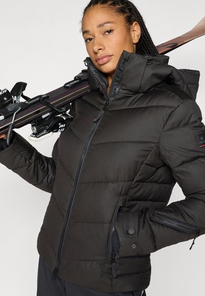 Μαύρο μπουφάν τύπου puffer με εφαρμοστό σχέδιο, με ψηλό γιακά, τσέπες με φερμουάρ και υφή με ραφές. Το μοντέλο κρατά σκι στους ώμους.