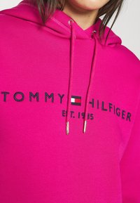 Růžová mikina s kapucí a šňůrkou, s černým logem a nápisem "TOMMY HILFIGER EST. 1985." Hladká textura a doplňky v odstínu zlata.