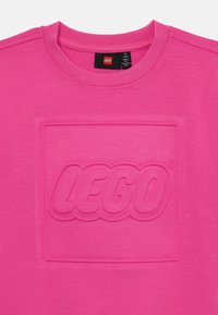 Fuchsia pink T-Shirt mit einem strukturierten LEGO-Logo in der Mitte, aus weicher Baumwolle mit Rundhalsausschnitt und kurzen Ärmeln.