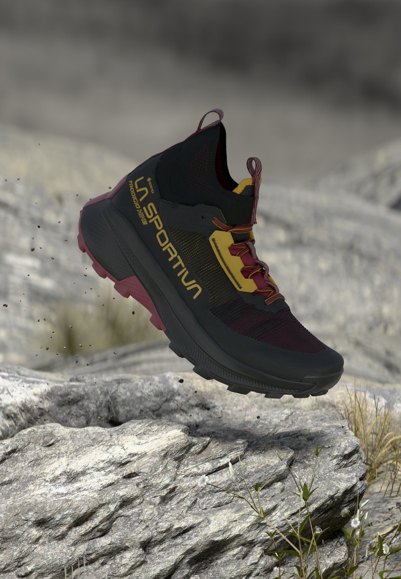 Černé a burgundské trailové běžecké boty s texturovaným svrškem, minimálním polstrováním mezi podrážkami a robustní podrážkou. Obsahuje žluté akcenty a tkaničky.