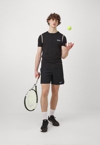 Camiseta atlética negra con logo, mangas cortas y rayas contrastantes en los hombros, combinada con pantalones cortos negros y una raqueta de tenis. Pelota de tenis en la mano.