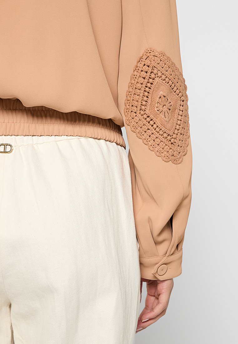 Blusa marrón claro con un detalle de ganchillo texturizado en la manga izquierda, cintura elástica y puños abotonados. Combinada con pantalones de color crema.