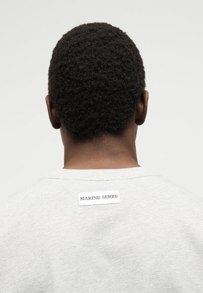 Άτομο με κοντά σγουρά μαλλιά γυρισμένο προς τα πίσω, που φοράει ένα ανοιχτό γκρι πουκάμισο με ετικέτα «MARINE SERRE» στο πάνω μέρος της πλάτης.
