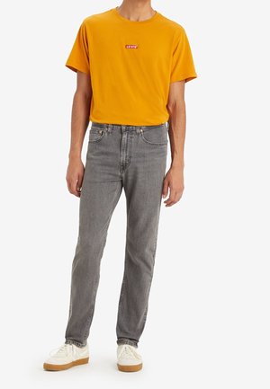 Man draagt een mosterdgele Levi’s T-shirt, grijze jeans en witte sneakers met rubberen zolen, staand tegen een witte achtergrond.