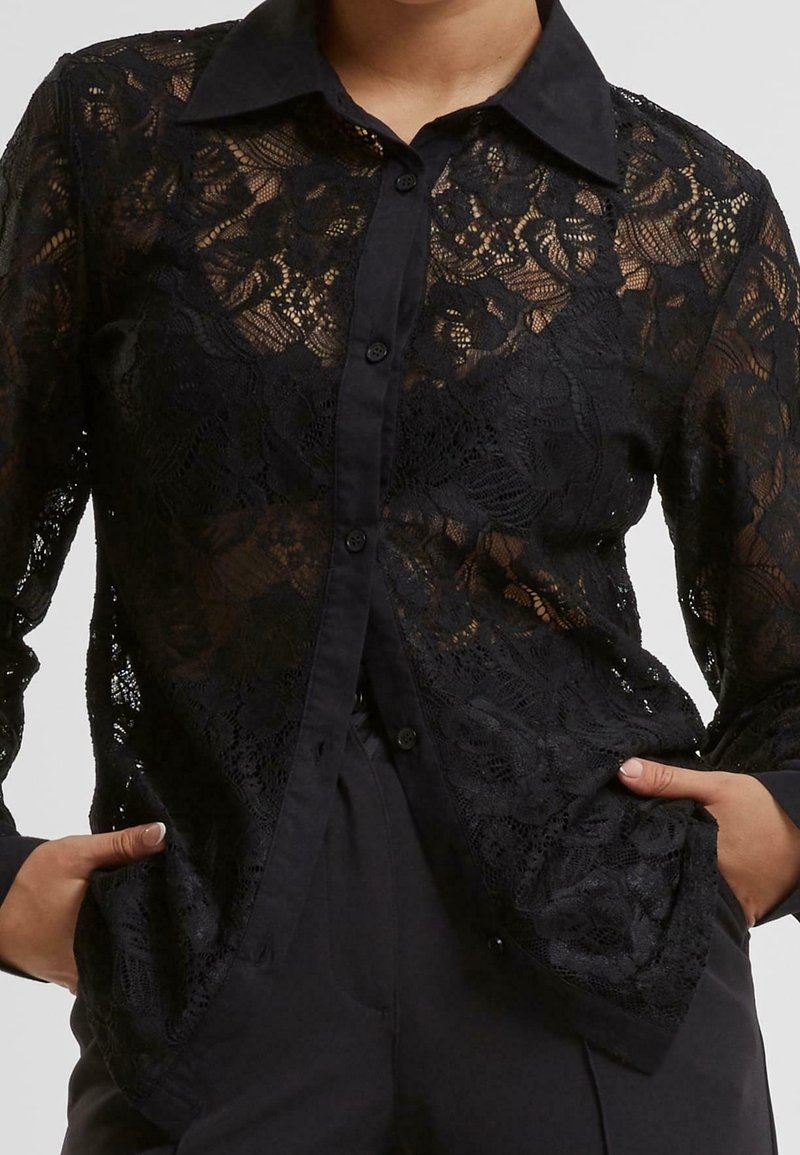 Chemise noire à manches longues en dentelle florale, boutonnée, portée avec les mains dans les poches, montrant un tissu transparent et un col et des poignets solides.