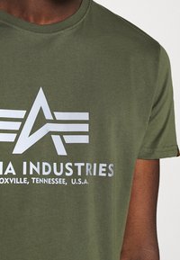Olivově zelené bavlněné tričko s kulatým výstřihem. Obsahuje bílé logo s abstraktním designem a textem "MA INDUSTRIES, KNOXVILLE, TENNESSEE, U.S.A."