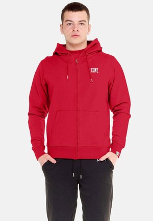 Jonge man staat met handen in de zakken, draagt een rode hoodie met rits en zwarte joggingbroek, kijkt recht vooruit tegen een effen achtergrond.