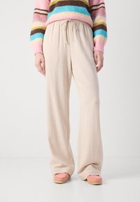 Pantaloni bej cu picioare largi, cu talie elastică și șnur, confecționați dintr-un material moale. Asortați cu pantofi roz slip-on, cu o talpă texturată.