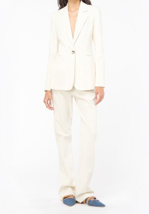 Kvinde iført en tætsiddende off-white blazer og matchende bukser med spidse blå sko med brune remme og nitter.