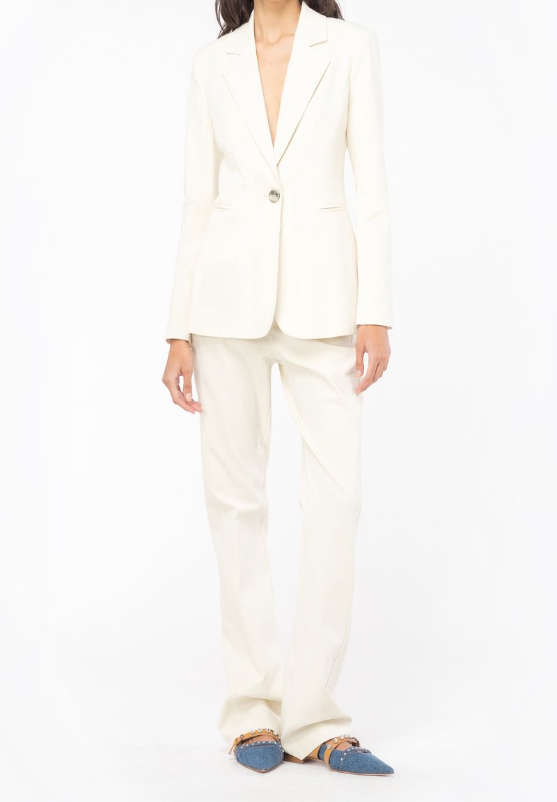 Femme portant un blazer ajusté écru et un pantalon assorti avec des chaussures pointues bleues ornées de sangles camel et de clous.