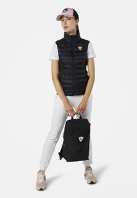 Schwarze, puffy Weste mit hohem Kragen, weißes T-Shirt, hellgraue Jogginghose und schwarzer Rucksack mit einem dreieckigen Logo sowie einer pinken Kappe.
