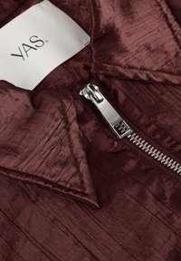 Close-up van een metalen rits op een getextureerde diepe bordeauxrode stof met een wit kledinglabel met de tekst "YAS."