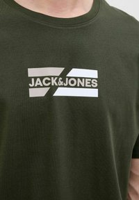 Oliven grøn bomulds t-shirt med et stort grafik i beige og hvid samt 'JACK & JONES' tekst, rund halsudskæring og korte ærmer.