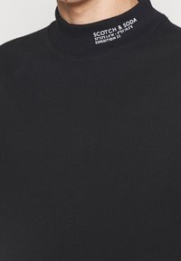 Sudadera negra de cuello alto hecha de algodón suave con texto blanco de la marca en el cuello. Diseño simple sin patrones ni acentos.
