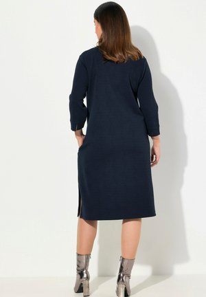 Navyblaues Kleid mit langen Ärmeln und seitlichen Schlitzen, aus strukturiertem Stoff, kombiniert mit metallischen Knöchelstiefeln.