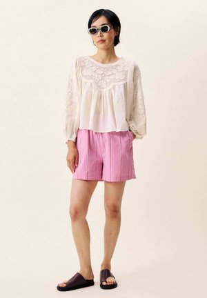 Femme portant un chemisier en dentelle blanche, un short plissé rose, des sandales plates foncées et des lunettes de soleil blanches, debout avec les mains dans les poches.