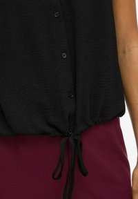 Črna srajca z gumbnim zapiranjem in nabranim robom, ki ima zavezan vrvico. Teksturovana tkanina se nežno spušča preko burgundskih hlač.