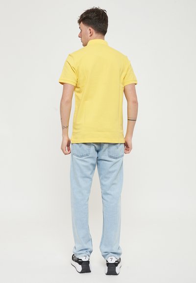 Polo giallo con colletto, abbinato a jeans in denim azzurri chiari. Le scarpe sono nere con accenti bianchi. Design semplice, vestibilità casual.