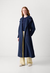 Long manteau bleu marine à double boutonnage, avec un col large et une coupe décontractée, associé à un pantalon jaune clair et des chaussures blanches.