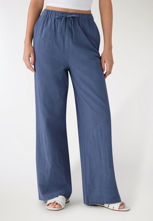 Donna che indossa pantaloni blu larghi con coulisse e sandali bianchi, in piedi con le mani rilassate lungo i fianchi su sfondo bianco.