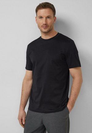 Mężczyzna ubrany w prostą czarną koszulkę z krótkim rękawem i szare spodnie, stojący na tle jasnoszarej ściany z jedną ręką w kieszeni.