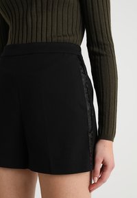 Svarta shorts med en strukturerad sidodekor, i kombination med en mörkgrön ribbad tröja. Shortsen har en skräddarsydd design och en åtsittande midja.