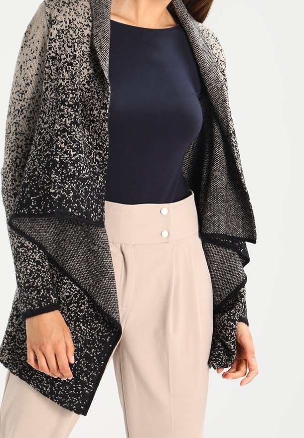 Femme portant un cardigan à motif noir et beige ouvert sur le devant, par-dessus un haut bleu marine, associé à un pantalon beige taille haute avec double bouton.