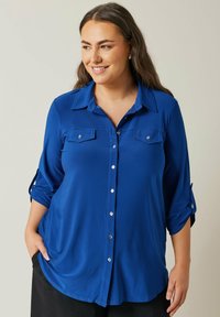 Camicia blu con bottoni, maniche lunghe arrotolate a tre quarti. Presenta due tasche sul petto e bottoni argentati lucidi. Tessuto dalla superficie liscia.