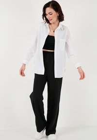 White button-up shirt over a black crop top, paired with loose black trousers and white sneakers. The shirt has a single chest pocket.