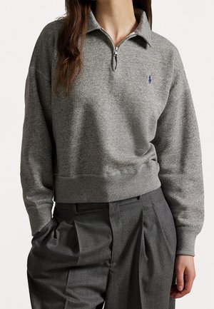 Vrouw die een grijs vest met ritssluiting en lange mouwen draagt, gecombineerd met donkergrijze geplooide broek, staand tegen een effen achtergrond.