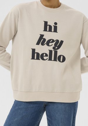 Beige Sweatshirt mit schwarzem Text "hi hey hello", getragen mit blauen Jeans, oben über dem Hals und den Knien abgeschnitten, Hände in die Hüften gestützt.