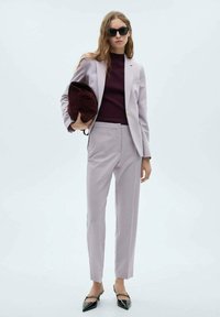 Helllila Anzug mit einer taillierten Jacke und geraden Hosen, kombiniert mit einem burgunderfarbenen Oberteil und einer schwarzen Handtasche. Spitz zulaufende schwarze Absätze runden den Look ab.